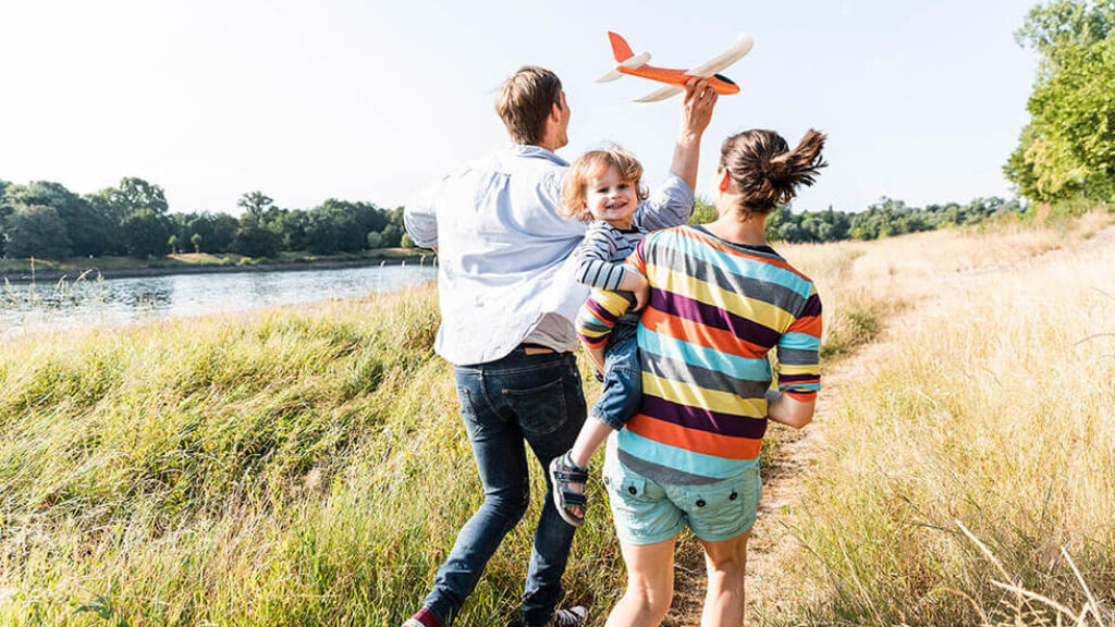 Famiglia corre nei campi. Padre tiene un aeroplano in mano e il bambino sorride felice in braccio alla mamma